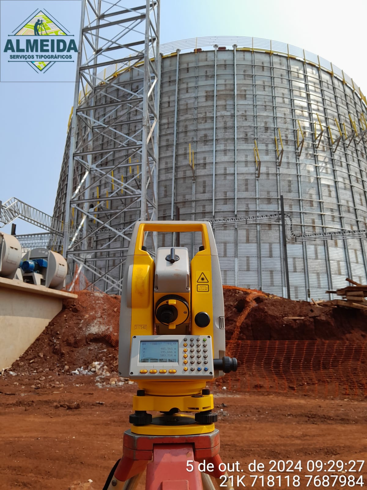 Equipamento topográfico diante de estrutura industrial metálica em obra.
